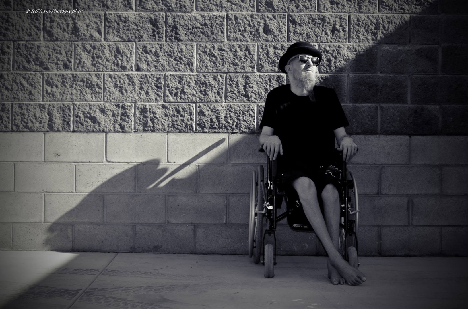Christopher Cox, a bearded man in a hat and sunglasses, seated in a wheelchair against a brick wall.